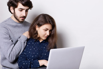 Young couple spending nice time together using high-speed internet connection on laptop. A man with trendy hairstyle and beard standing behind his girlfriend watching something on notebook