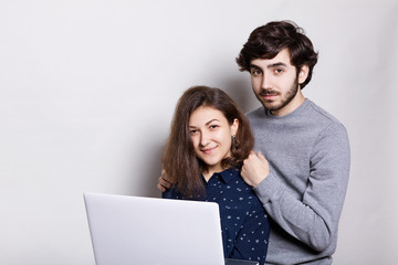 Fototapeta premium A happy man and woman standing sideways with laptop looking directly into camera isolated over white background. Stylish guy with beard standing behind his girlfriend enjoing modern technologies.