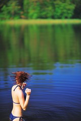Fototapeta premium Young girl in bikini, standing in the river and shakes his head, flicking water out of your hair. Water drops fly in air, creating a beautiful pattern. Scenic summer background.