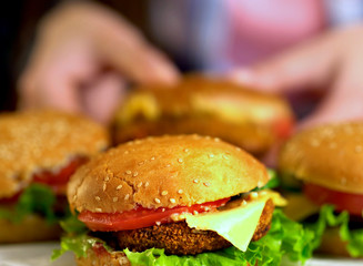 Hamburger fast food with ham on wooden board . Burger with fresh vegetables and cheese slice on blurred background. Piece of cheese hanging from sandwich In blur. Female hand reaches for cheeseburger