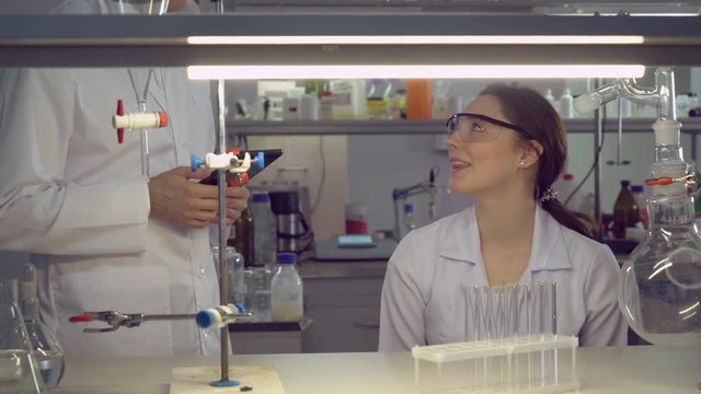 Attractive smiling woman sitting at the working place with different medical equipment flasks and glass bulb. researchers discussing experiments. woman explain with happy smile.