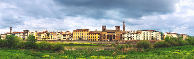 Fototapeta premium Beautiful panoramic view of Florence riverside, Arno River, National Library in the middle, green grass in foreground and majestic powerful cloudy sky
