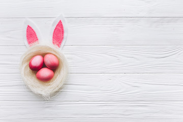 Nest with three pink Easter eggs and bunny ears on the wooden white background.