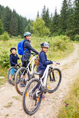 Obraz premium Young girl with her mother and small brother enjoying cycling in forest. Family riding bike concept background