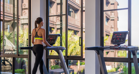 Fototapeta na wymiar Horizontal shot of woman jogging on treadmill at health sport club at luxury resort. Female working out at a gym running on a treadmill with a great summer view at spa facility at hotel during summer
