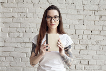 Pretty hipster tattooed woman poses in white t-shirt, isolated on white brick wall, Hold a book in hand, space for design layout