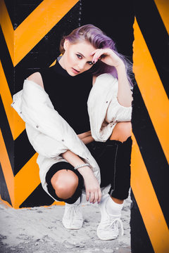 A beautiful girl with purple hair walks the streets of the city. Wearing a jacket, torn jeans. Sits in a parking lot between posts