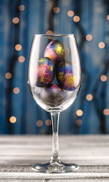 Easter Eggs Painted In Space In A Luxurious Wine Glass On A Wooden Table In The Background Of Lights