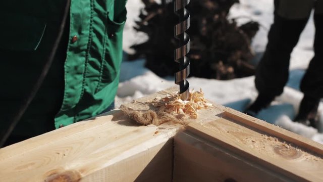 Close up drilling hole in two wooden blocks corner connection creating sawdust outside in winter, snow on background
