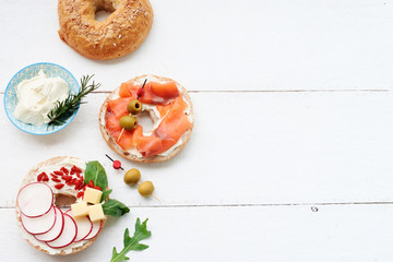 Assorted bagels with cream cheese, italian ham, cherry tomatoes, rocket salad, radish, cheddar, salmon and olives over white background. Copy space.
