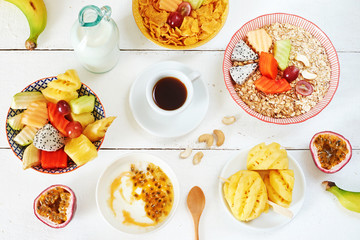 Assorted breakfast with exotic fruits. Oats, cornflakes, yoghurt, pineapple, passionfruit, papaya, banana, guava, dragon fruit, melon, grapes. Black coffee and milk on a white table. 