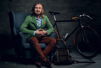 A man dressed in a green jacket sits on a chair with single speed bicycle on background.