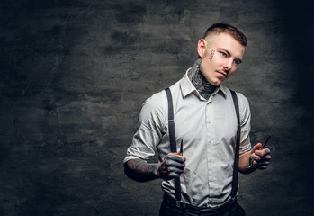 Portrait of tattooed male barber holds sharp scissors and a blade.