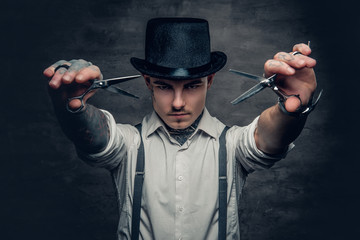Portrait of tattooed male barber holds sharp scissors and a blade.