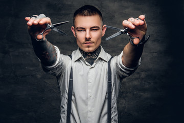 Portrait of tattooed male barber holds sharp scissors and a blade.