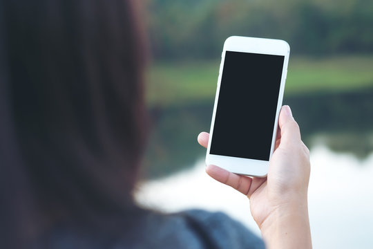 Mockup image of a woman using white smart phone with blank black screen at outdoor and lake nature background