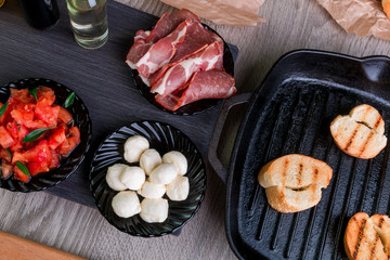 Toast bread on grill pan near plates with mozzarella, ham, tomato. Bruschetta ingredient. Top view.