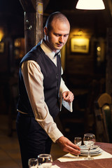 Waiter served table in the restaurant before the feast,Waiter in uniform waiting an order,Waiter with a white towel on his hand,Confident waiter,A pub.Restaurant.Classic.Evening.European restaurant