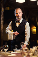 Waiter served table in the restaurant before the feast,Waiter in uniform waiting an order,Waiter with a white towel on his hand,Confident waiter,A pub.Restaurant.Classic.Evening.European restaurant