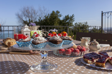 Chocolate muffins with cherry on a dish.  Sliced cheesecake in the background, with blue sky. Breakfast time