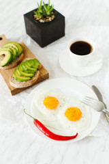 Morning breakfast, table setting. On a white plate are two fried eggs, on a wooden board sliced avocado, a cup of coffee and a vase with a plant. White background, daylight, vertical image.