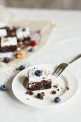 Table setting, chocolate dessert brownie cake with berries on a white plate on a table covered with a linen tablecloth. The second part of the cake on the parchment paper. Vertical shot, daylight.
