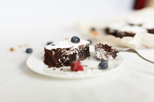 Table setting, chocolate dessert brownie cake with berries on a white plate on a table covered with a linen tablecloth. The second part of the cake on the parchment paper. Horizontal image, daylight.