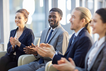 Multi-ethnic group of smiling business people sitting in row in modern glass hall and clapping, focus on cheerful African businessman looking at camera