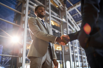 Low angle portrait of two business partners in handshake: smiling African -American businessman shaking hands with Caucasian colleague in hall of modern glass office building at night time