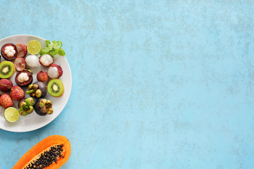 Fresh tropical fruits from Thailand over blue background. Cut in half papaya. Lychee, mangosteen, kiwi and lime on a plate. Healthy summer breakfast.