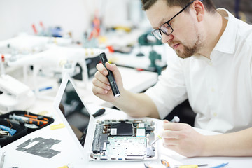 Side view portrait of man opening disassembled computer and using flashlight to inspect broken...