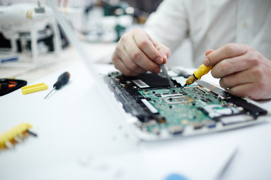 Closeup shot of unrecognizable man fixing circuit board in laptop using screwdriver and different tools on table in workshop