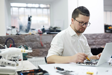 Portrait of modern man wearing glasses and creative haircut busy working with electronics in workshop: disassembling laptop and drones with different tools and electronic devices on table