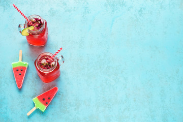 Fototapeta na wymiar Two portions of homemade watermelon lemonade in mason jar with red striped straw and watermelon popsicles on a blue background