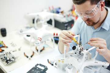 Man in protective glasses attaching action camera to quadcopter drone on table  with different tools in modern workshop