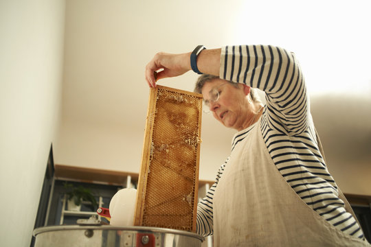 Senior female beekeeper scraping honeycomb into kitchen saucepan