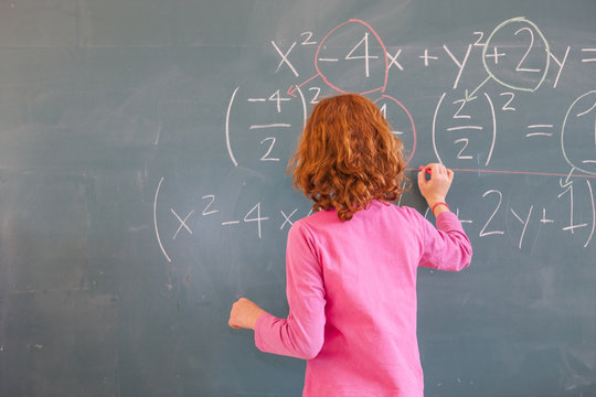Rear view of primary schoolgirl answering equation on classroom blackboard