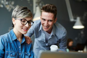 Smiling woman with stylish haircut finishing project on laptop while her handsome coworker looking...