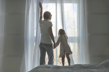 Woman and little girl, wake up and open the curtains in the morning, back view