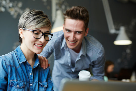 Smiling woman with stylish haircut finishing project on laptop while her handsome coworker looking at computer screen and patting her shoulder as sign of approval
