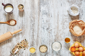 Baking background. Wholegrain flour, eggs in a carton tray, oat flakes, grains and seeds, milk, olive oil, rolling pin, wooden spoon over light rustic table. Space for text.