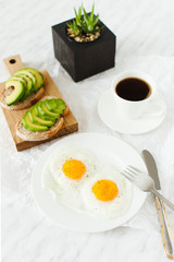 Morning breakfast, table setting. On a white plate are two fried eggs, on a wooden board sliced avocado, a cup of coffee and a vase with a plant. White background, daylight, vertical image.