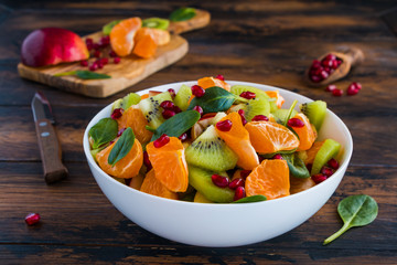 Healthy dietary fruit salad with fresh kiwi, tangerines, apples, spinach and pomegranate seeds in a white bowl on the wooden rustic table, a cutting board, a knife