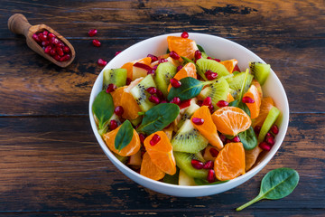 Vitamin fruit salad with fresh apples, tangerines, kiwi, spinach and pomegranate seeds. White bowl on the wooden rustic table.