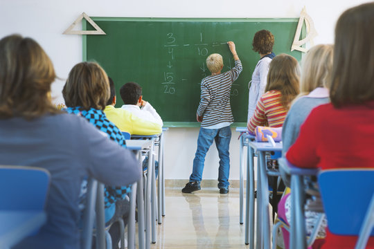Boy writing sum on blackboard in front of class, rear view