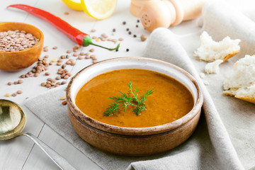 Delicious soup made of lentils in a rustic clay bowl served on a canvas tablecloth. Healthy vegetarian meal with bread, lemon and chili pepper on a table.