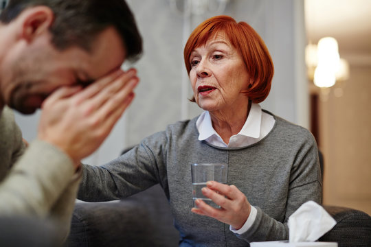 Portrait of pretty mature psychologist trying to comfort her crying middle-aged patient and offering him glass of water, box with paper handkerchiefs standing on table