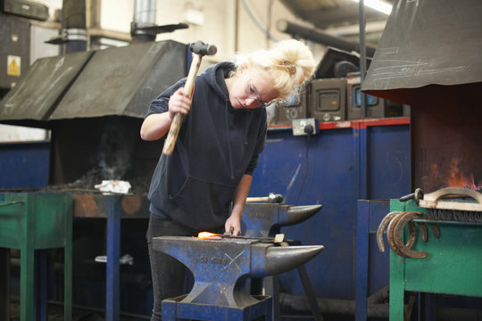 Young female trainee blacksmith hammering horseshoe on workshop anvil - Powered by Adobe