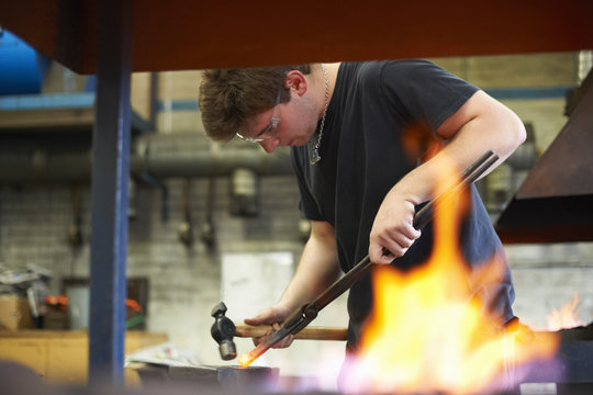 Young male trainee blacksmith hammering red hot metal on workshop anvil