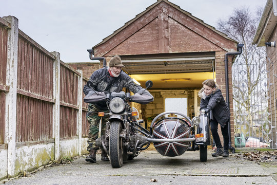 Senior male motorcyclist and grandson pushing motorcycle and sidecar from garage